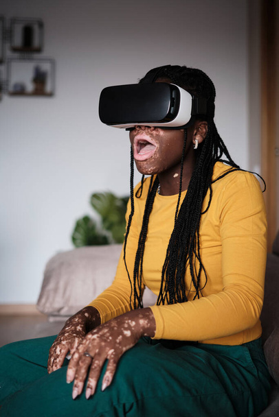 Genç Afro-Amerikalı kadın, günlük kıyafetleriyle sanal gerçeklik gözlüklerini keşfediyor, evde otururken ağzı açık hayret içinde. - Fotoğraf, Görsel