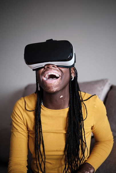 Fröhliche schwarze Frau im gelben Pullover mit Afro-Zöpfen erlebt Cyberspace in Virtual-Reality-Brille zu Hause - Foto, Bild