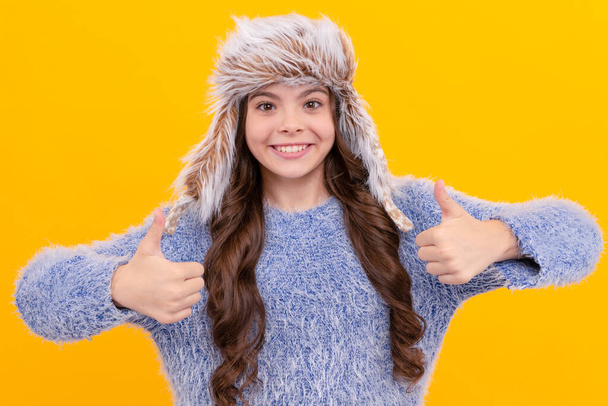 winter fashion. thumb up. happy kid with curly hair in earflap hat. female fashion model. teen girl in knitwear on yellow background. portrait of child wearing warm clothes. express positive emotion. - Photo, Image