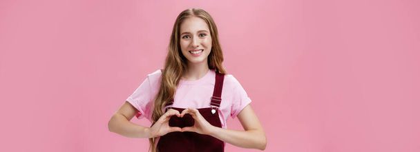 Girl loves family. Kind charming young woman in overalls with small tattoo on arm showing heart gesture over body and smiling lovely at camera expressing tender and cute attitude over pink wall. - Photo, Image