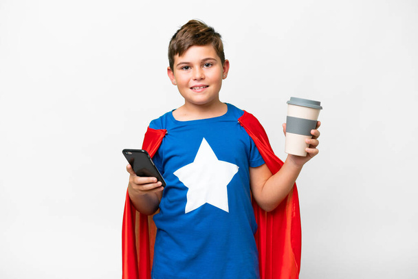 Super Hero caucasian kid over isolated white background holding coffee to take away and a mobile - Foto, immagini
