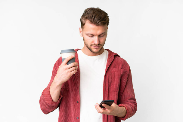 Young handsome caucasian man over isolated white background holding coffee to take away and a mobile - Φωτογραφία, εικόνα