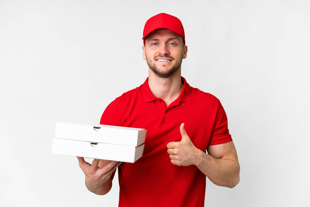 Pizzalieferant in Arbeitsuniform nimmt Pizzakartons vor isoliertem weißen Hintergrund auf und macht eine Daumen-hoch-Geste - Foto, Bild