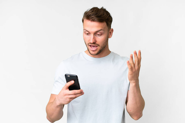 Young handsome caucasian man over isolated white background looking at the camera while using the mobile with surprised expression - Фото, зображення