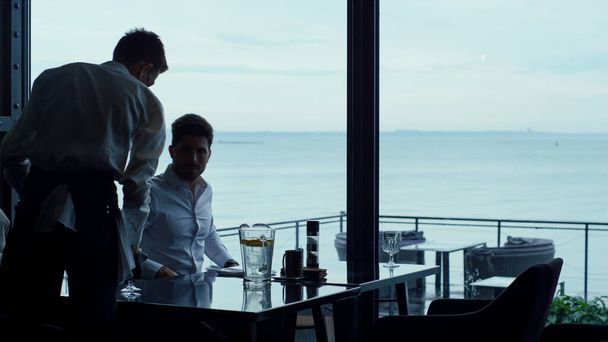 Elegant waiter silhouette serving successful businessman customer in seascape restaurant. Handsome man resting weekend lunch in lounge hotel bar. Cafe at covid 19 concept. Luxury lifestyle concept. - Fotografie, Obrázek