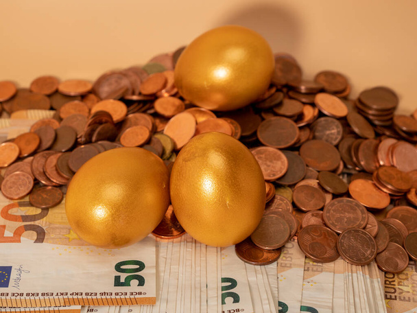 Gouden eieren op geld. Gouden kippeneieren op eurobankbiljetten en -munten. Rijkdom symbool. Sparen en beleggen. - Foto, afbeelding