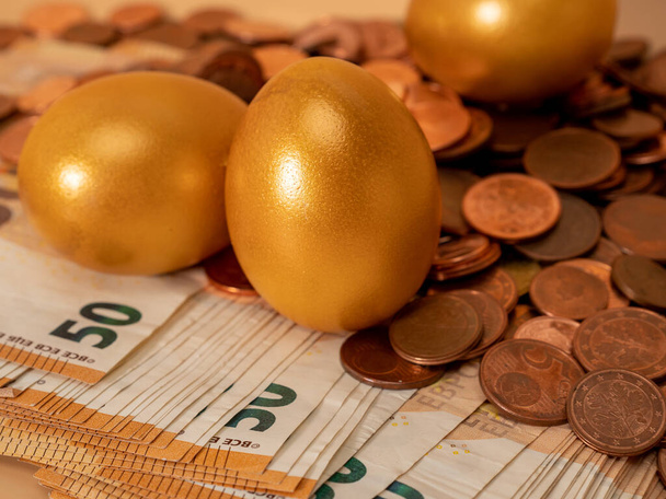 Golden eggs on money. Golden chicken eggs on euro banknotes and coins. Wealth symbol. Savings and investment. - Photo, Image