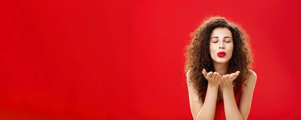 Tender and gentle stylish caucasian girl with curly hairstyle. and red lipstick bending towards camera with slight smile closed eyes and palms near folded lips blowing kiss at camera romantically. - Photo, Image