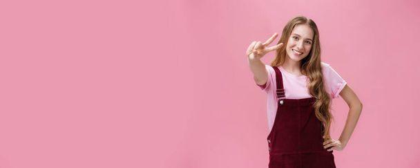 Lebensstil. Charismatische junge Teenagerin in Cordhosen posiert freudig und energisch vor rosa Hintergrund und zeigt ein Friedenszeichen in die Kamera, hält die Hand auf der Taille in selbstbewusster Pose und lächelt - Foto, Bild