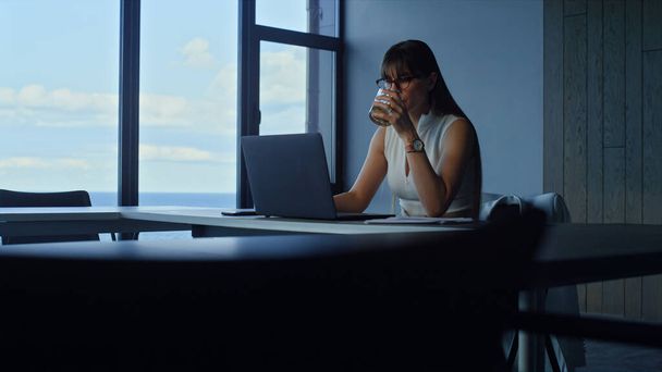 Focused businesswoman drinking water in empty office room. Ceo overworking alone creating corporate report surfing internet in glass office. Successful financial analyst look using laptop computer - Foto, afbeelding