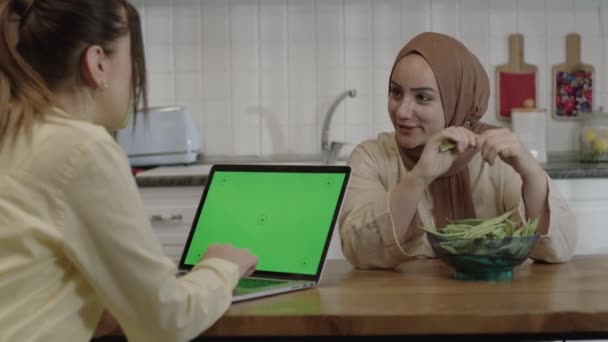A woman dressed in a hijab, preparing food at the kitchen table, chats with a woman doing her job on a green screen computer. Portrait of housewife and businesswoman friendship. - Záběry, video