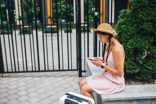 Young slender woman is sitting on a bench with mobile phone, gray suitcase standing by legs, side photo - 写真・画像