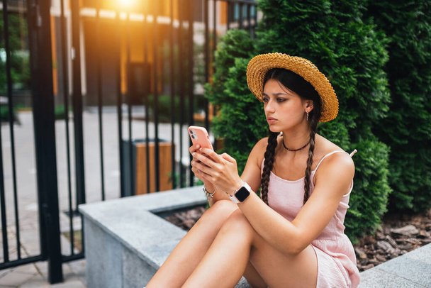 Giovane donna snella è seduta su una panchina con telefono cellulare, valigia grigia in piedi da gambe, foto laterale - Foto, immagini
