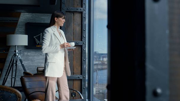 Thinking woman drinking coffee at panorama window. Focused manager having break in office alone. Successful businesswoman making decision at sea view. Thoughtful brunette lady resting with espresso - Foto, imagen