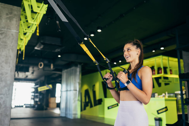 Junge athletische Frau mit perfektem Körper in Sportbekleidung bei Übungen mit Trageschlaufen im Fitnessstudio - Foto, Bild