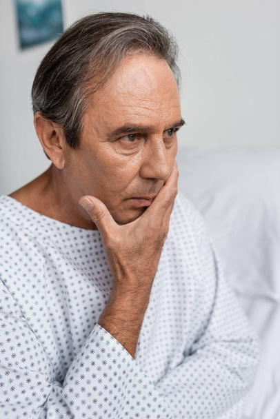 Disappointed elderly man in patient gown looking away in hospital ward - Foto, imagen