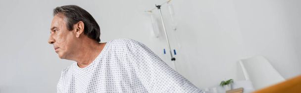 Vista lateral do homem idoso em vestido de paciente olhando para longe na enfermaria do hospital, banner - Foto, Imagem