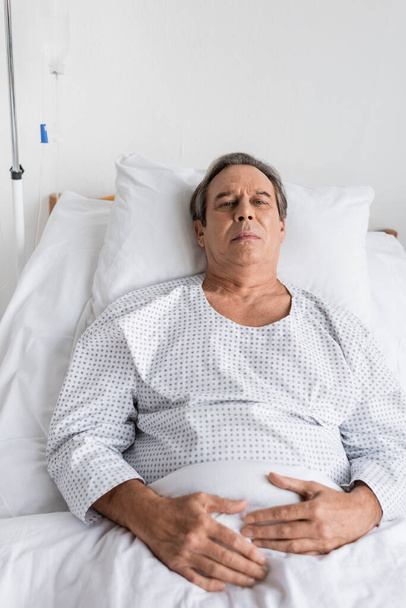 ziek grijs harige patiënt in toga kijken naar camera terwijl liggend op bed in kliniek - Foto, afbeelding