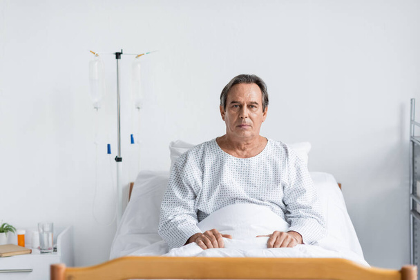 Seniorin blickt in Kamera, während sie auf Krankenhausstation im Bett sitzt - Foto, Bild