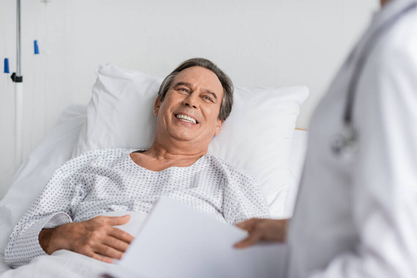 Cheerful patient in gown looking at blurred doctor with paper folder in hospital ward - Foto, Imagen