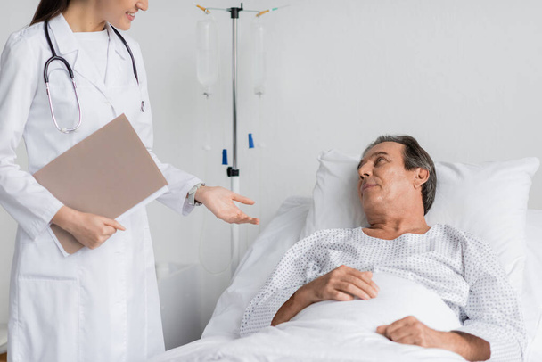Smiling doctor with paper folder talking to senior patient on bed in clinic - Foto, immagini