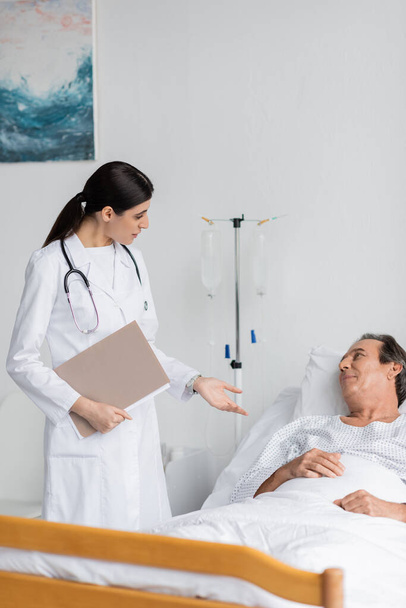 brunette doctor with paper folder talking to elderly patient in clinic - Photo, Image