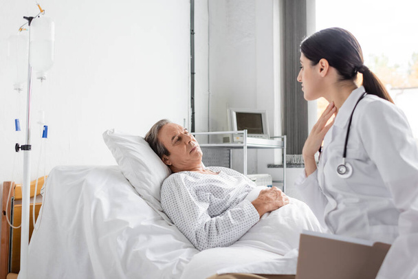 Doctor with paper folder touching neck while talking to senior patient in hospital ward  - Foto, Imagen
