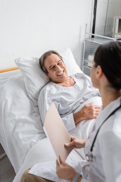 Positive senior patient looking at blurred doctor with paper folder in hospital ward  - Foto, Imagen