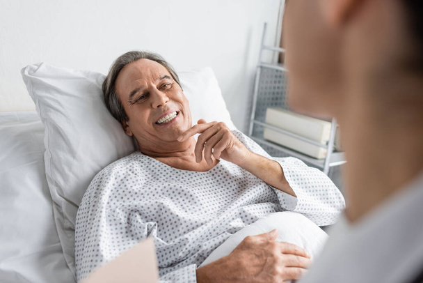 Cheerful elderly patient looking at blurred doctor in hospital ward  - Photo, image