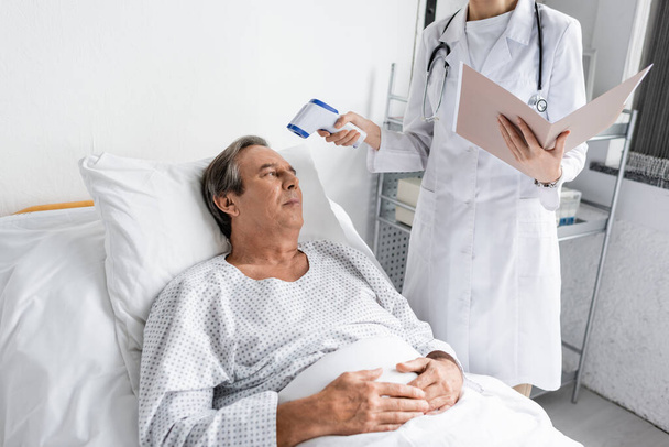 Doutor com pasta de papel segurando pirômetro perto do paciente sênior no hospital  - Foto, Imagem
