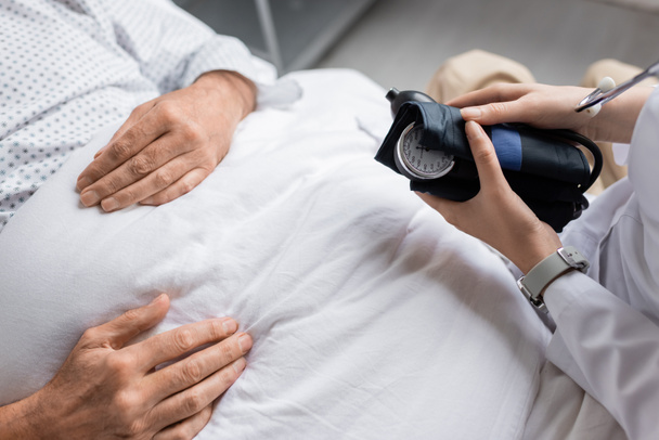 Обрезанный вид врача, держащего тонометр рядом с больным пациентом на больничной койке  - Фото, изображение