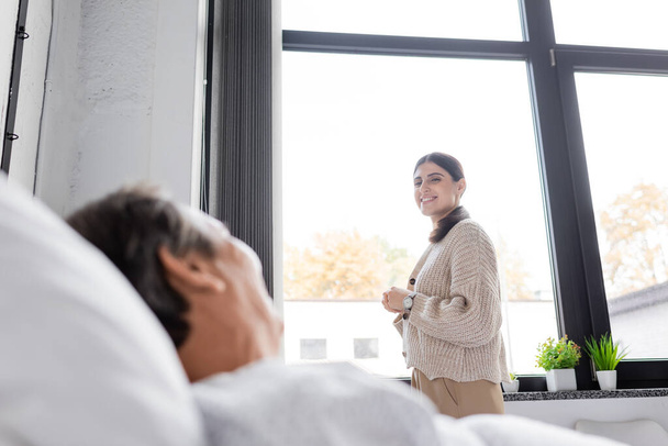 Uśmiechnięta kobieta patrząca na niewyraźnego ojca leżącego na szpitalnym łóżku  - Zdjęcie, obraz