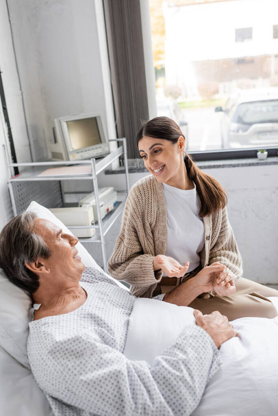Positive woman holding hand of father in patient gown and talking in hospital ward  - Foto, Imagen