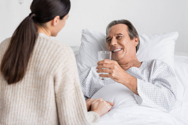 Усміхнений старший пацієнт тримає склянку води біля розмитої дочки в клініці
  - Фото, зображення
