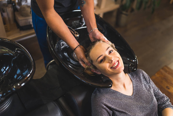 Visão de alto ângulo de sorrir mulher relaxada tendo seu cabelo lavado e massageado por trabalhador cabeleireiro irreconhecível. Lavar o cabelo no salão de cabeleireiro. Foto de alta qualidade - Foto, Imagem