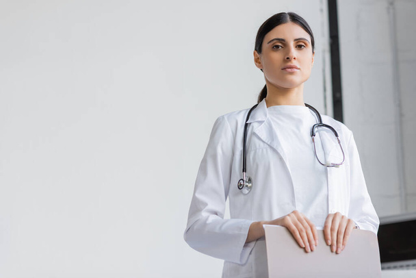 Tiefansicht des Arztes im weißen Kittel mit Papiermappe und Blick in die Kamera in der Klinik  - Foto, Bild