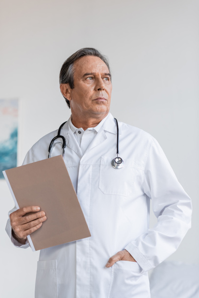 Oberarzt hält Papiermappe in der Hand und schaut in Klinik weg  - Foto, Bild