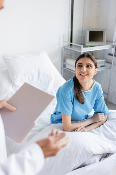 Χαμογελώντας ασθενής κοιτάζοντας το γιατρό με το φάκελο χαρτί στην κλινική  - Φωτογραφία, εικόνα