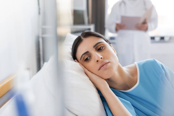 Dissatisfied patient lying on bed near blurred doctor in hospital ward  - Фото, зображення