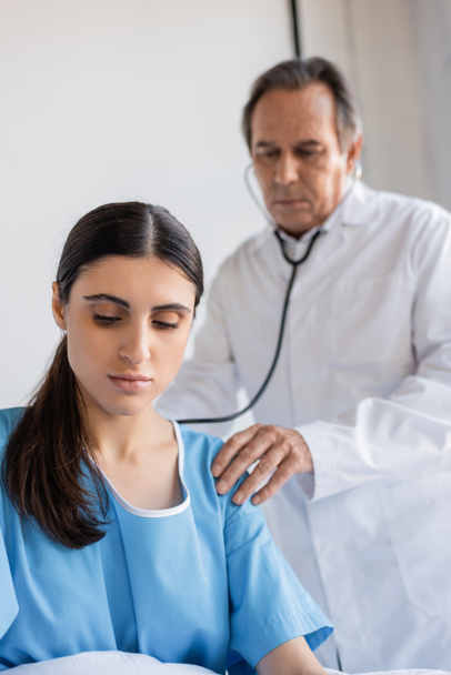 пациент в халате сидит рядом с размытым врачом со стетоскопом в клинике  - Фото, изображение