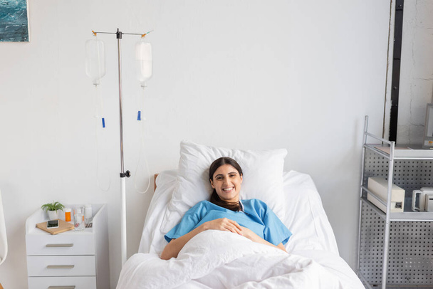 Vrolijke patiënt kijkt naar camera in de buurt van intraveneuze therapie in kliniek  - Foto, afbeelding