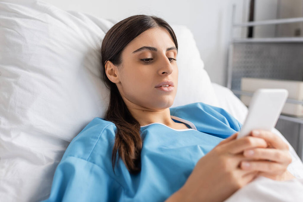 sick woman in patient gown using smartphone on bed in hospital  - Foto, Imagen