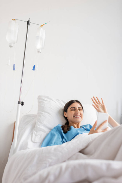Lachende patiënt met videogesprek op smartphone in de buurt van intraveneuze therapie in het ziekenhuis  - Foto, afbeelding