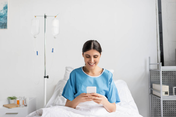 glückliche Frau im Patientenkleid lächelt, während sie das Smartphone auf dem Bett in der Klinik benutzt  - Foto, Bild