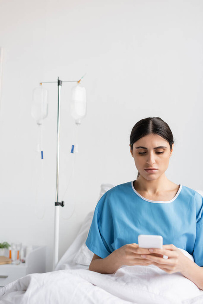 paziente bruna che usa il cellulare mentre è seduto sul letto in ospedale  - Foto, immagini
