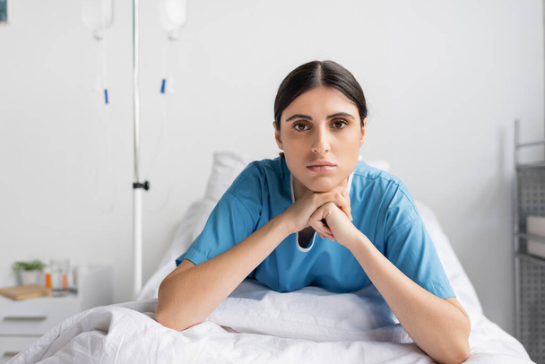 brünette Frau im Patientenkleid blickt in die Kamera, während sie in der Klinik auf dem Bett sitzt  - Foto, Bild