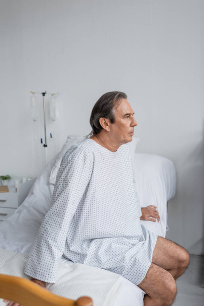 Γκρίζα μαλλιά ασθενή με φόρεμα κάθεται στο κρεβάτι στο νοσοκομείο  - Φωτογραφία, εικόνα