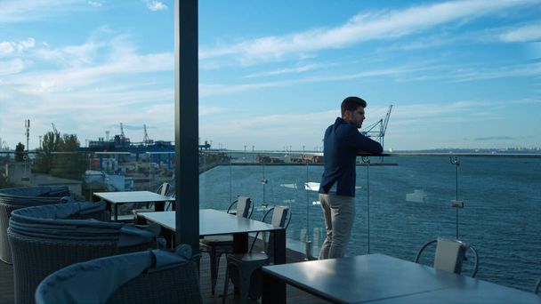 Красивый мужчина, отдыхающий на террасе в морском порту. Сосредоточенный бизнесмен думает о проблемах бизнеса, наслаждаясь спокойным утром в роскошном кафе. Привлекательный менеджер брюнеток с видом на океан, отдыхающий на балконе - Фото, изображение