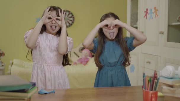 Portrait of happy little girls making funny faces, posing in their room at home. Girls making binoculars with fingers having fun together, showing glasses with hands, posing, looking at camera. - Imágenes, Vídeo