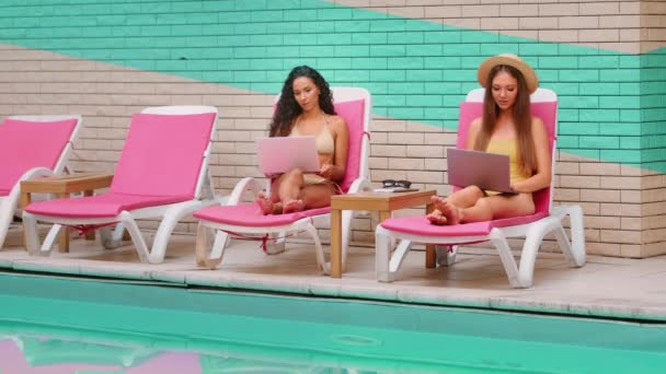 Two millennial girls Caucasian Latina women millennial relax on sun loungers by summer pool work remotely use laptop search media content booking hotel travel business freelance or distance education - Footage, Video